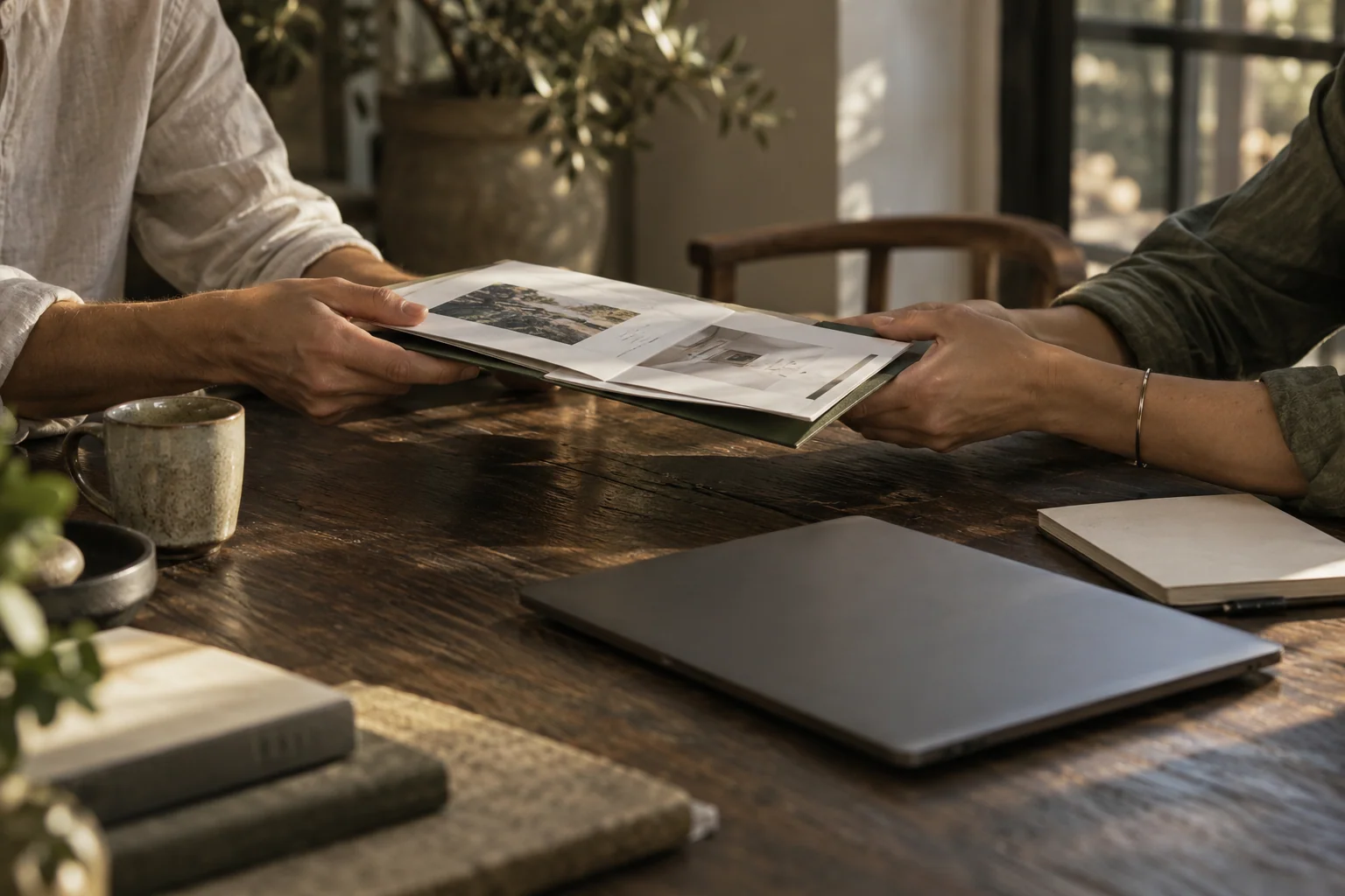 Two coworkers passing a project folder across a sunlit wooden table.